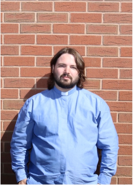 Revd Mark Wilson in a pale blue clerical shirt standing in front of a plain brick wall