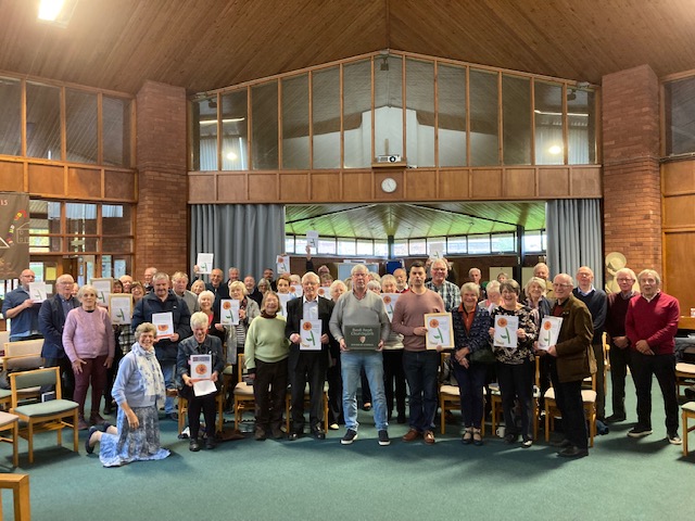 A group photo with around 50 people, many holding Churchyard Award certificates gathered inside the modern St Thomas Church, Doxey
